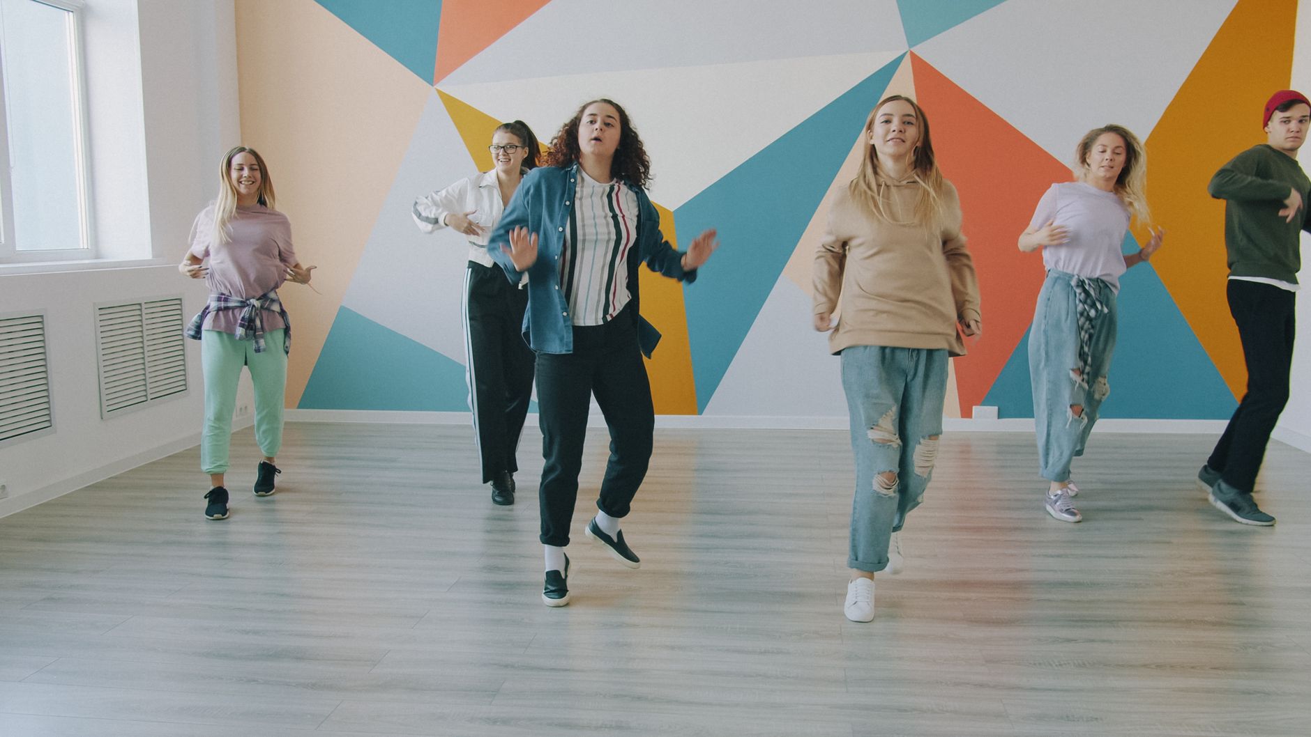 Joyful dance group practicing in a bright room with a geometric mural, expressing movement and happiness.